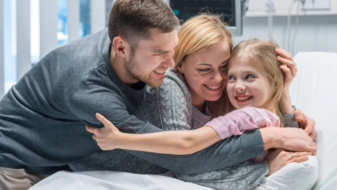 A family hugging their young daughter in her hospital bed.