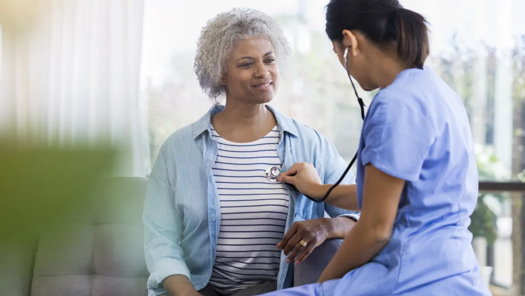 A female doctor is seated, listening to a senior woman's heart with a stethoscope.