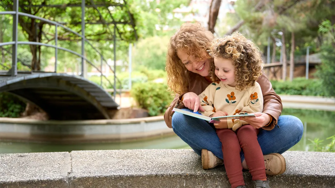 Mother and child reading a book together while sitting on a wall by a river.