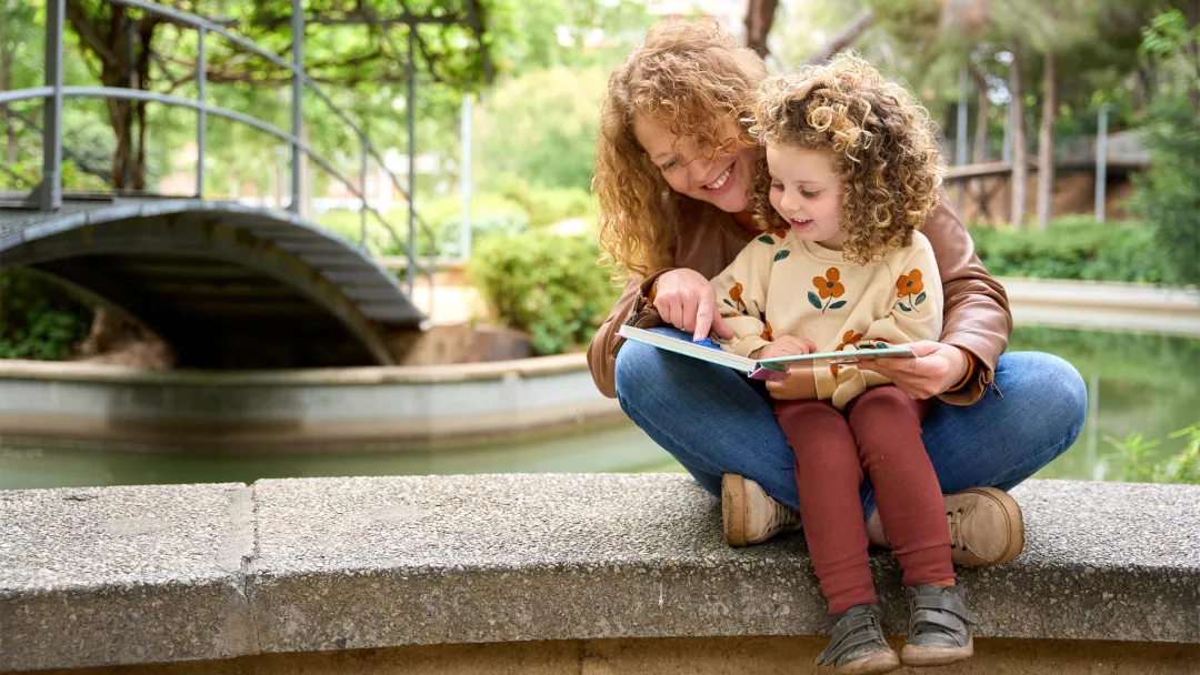 Mother and child reading a book together while sitting on a wall by a river.