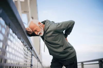 A senior man hunching over against a fence with his hand resting on his back in pain.