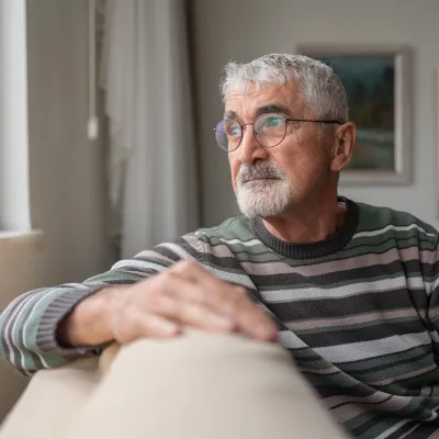 An Senior Man with gray hair and glasses is sitting on a beige couch, wearing a striped sweater.