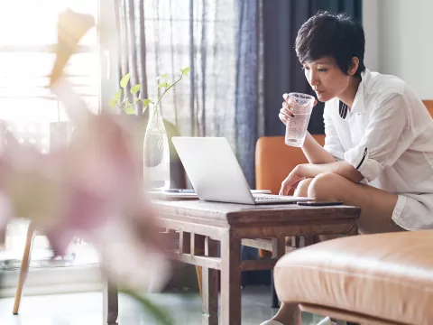 A woman drinks a cup of water while working on her laptop at home