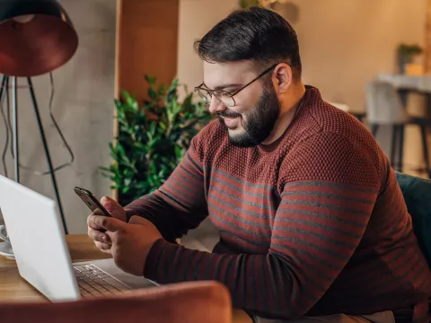 A Man Smiles as He Uses a Cellphone While Sitting at a Desk in front of a Laptop.