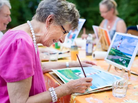 A Grandmother Paints a Watercolor Painting Based on an Example on a Tablet.