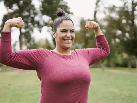 A Woman Flexes Her Muscles in a Park with a HUge Smile on Her Face.