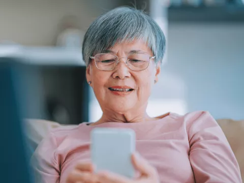 A Senior Woman Sits on the Couch at Home and Scrolls on Her Cell Phone
