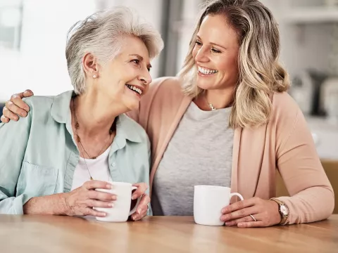 Adult woman with her arm around her mother, both smiling and holding white mugs.