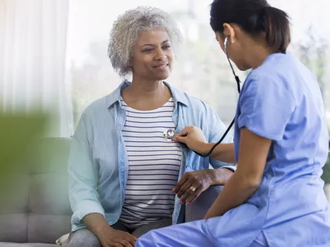 A female doctor is seated, listening to a senior woman's heart with a stethoscope.