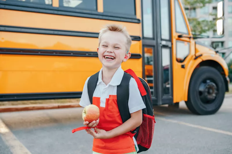A Small Boy Laughs as He Gets Off a School Bus with an Apple in Hand.