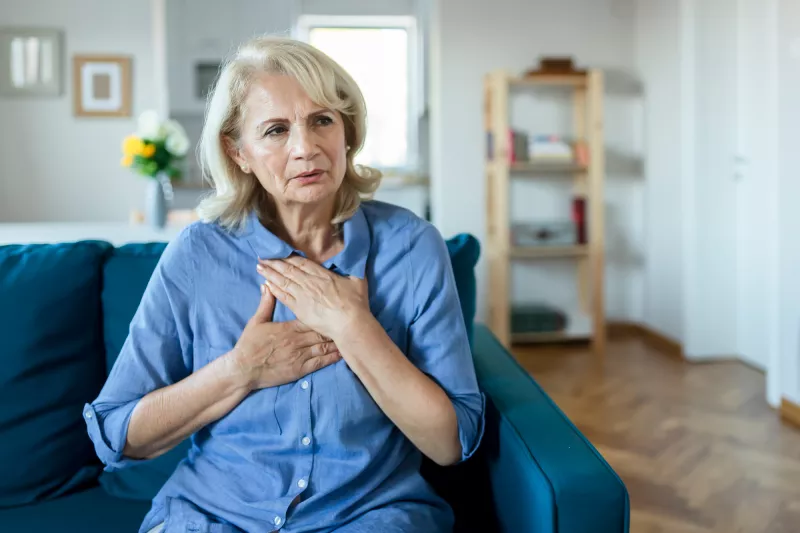 A Woman Sits in Her Living Room With Her Hands on Her Chest, Feeling Her Heartbeat.