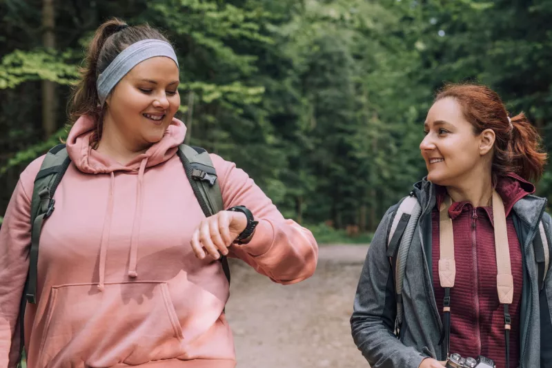 Two Women Go For a Hike Through the Woods as One of Them Checks Their Vitals on Their Smart Watch.