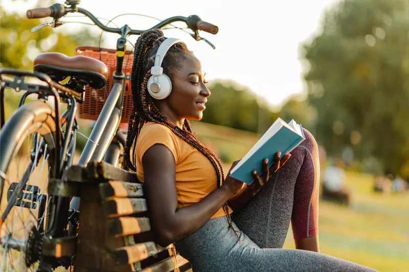 A Woman Takes a Break From Riding Her Bike By Sitting on a Park Bench and Reading a Book.