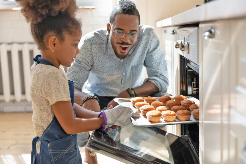 A Little Girl Pulls Muffins From an Oven While Her Father Watches