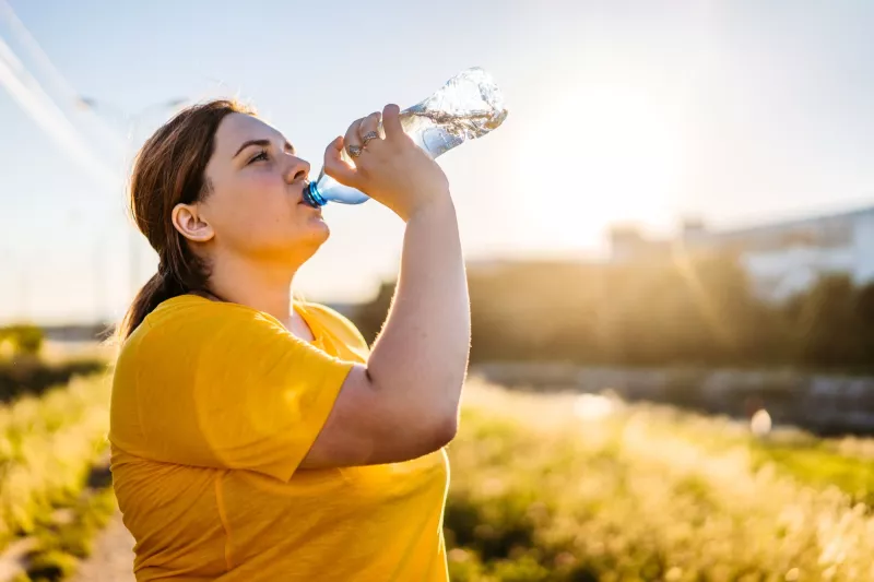 A woman drinking a bottle of water on a sunny day, surrounded by yellow flowers.
