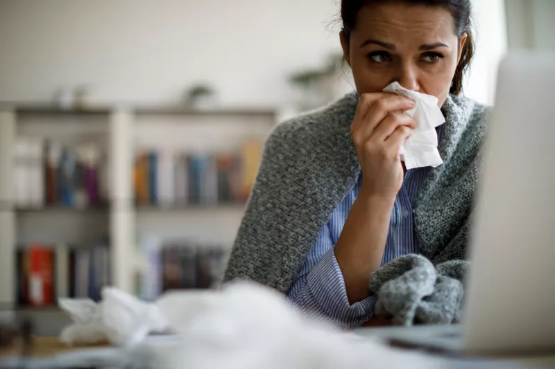 A Woman Surfs the Internet on a Laptop as she Touches Her Nose With a Tissue.