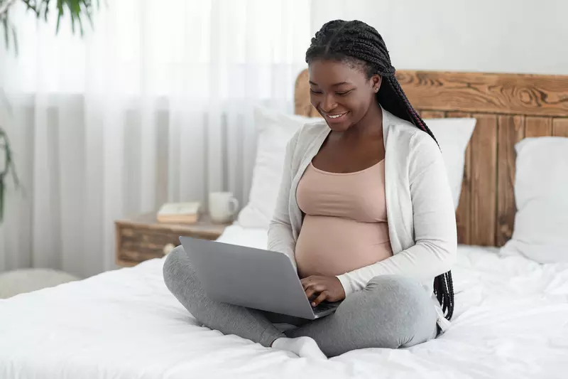 A Smiling Pregnant Woman Sits on Her Bed, in Her Home, Using a Laptop 