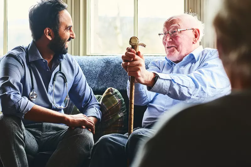 A doctor, sitting on a sofa with a senior man holding a cane, making a home visit.
