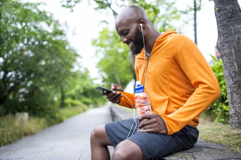 An adult man in an orange hoodie, sitting on a bench outside, looking at his phone and hydrating with a bottle of water.