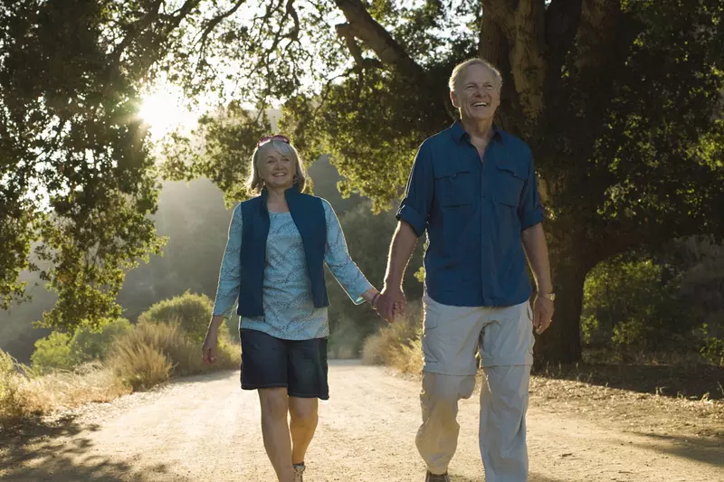 An older Caucasian couple takes an evening walk on a trail.