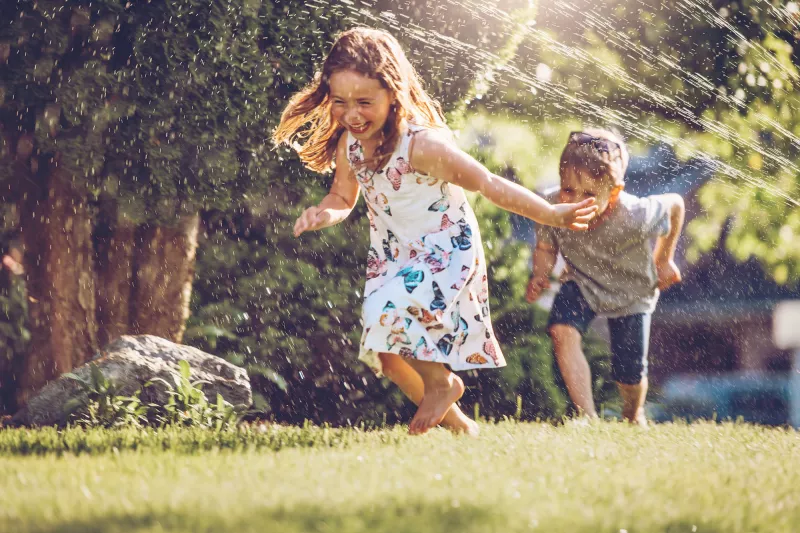 Two children playing in the yard with sprinklers.