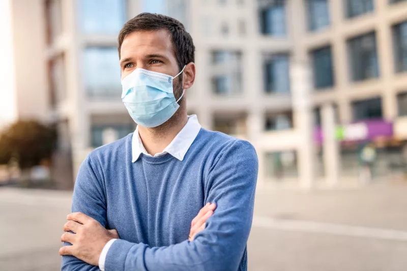 A man with his arms crossed standing outdoors wearing a mask.