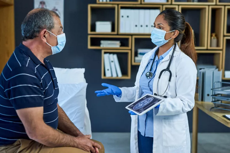 A masked doctor talking to her masked patient with a tablet on her hand