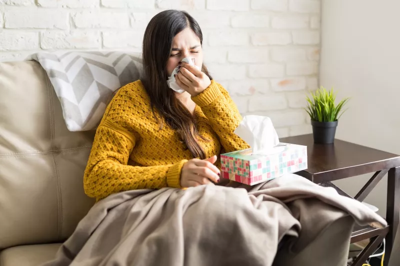 A young woman sitting on her couch sneezing.