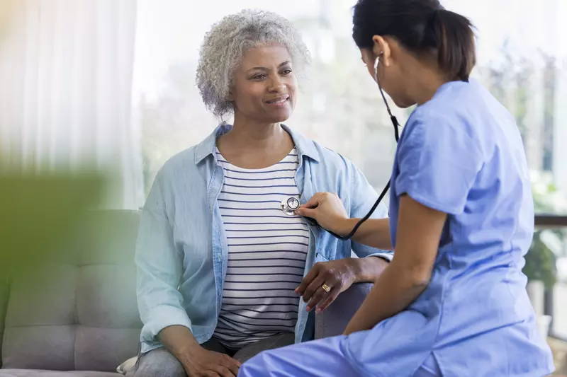 A female doctor is seated, listening to a senior woman's heart with a stethoscope.