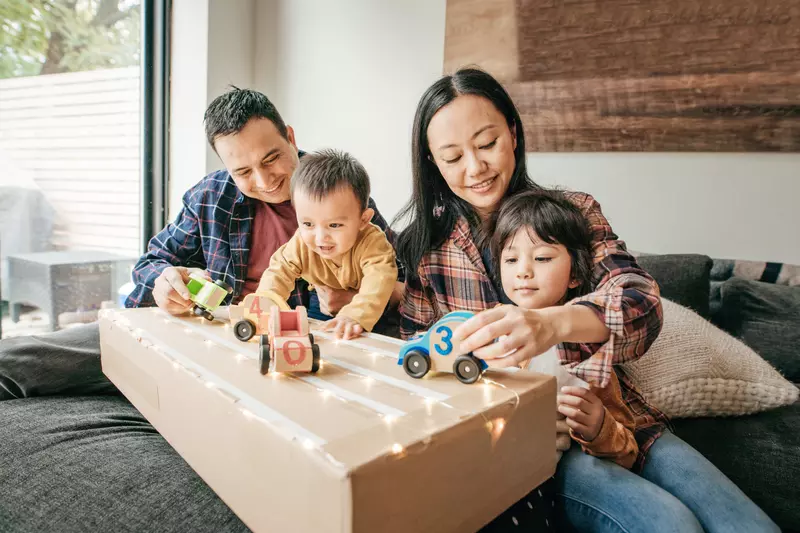 Parents and two children playing with wooden cars on top of a cardboard box while sitting on the couch at home.