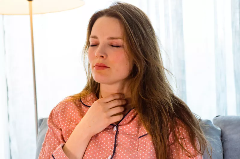 Woman touching her sour throat while sitting at home while wearing pajamas.