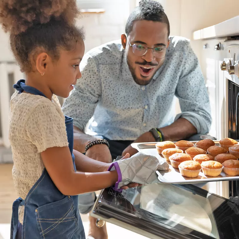 A Little Girl Pulls Muffins From an Oven While Her Father Watches