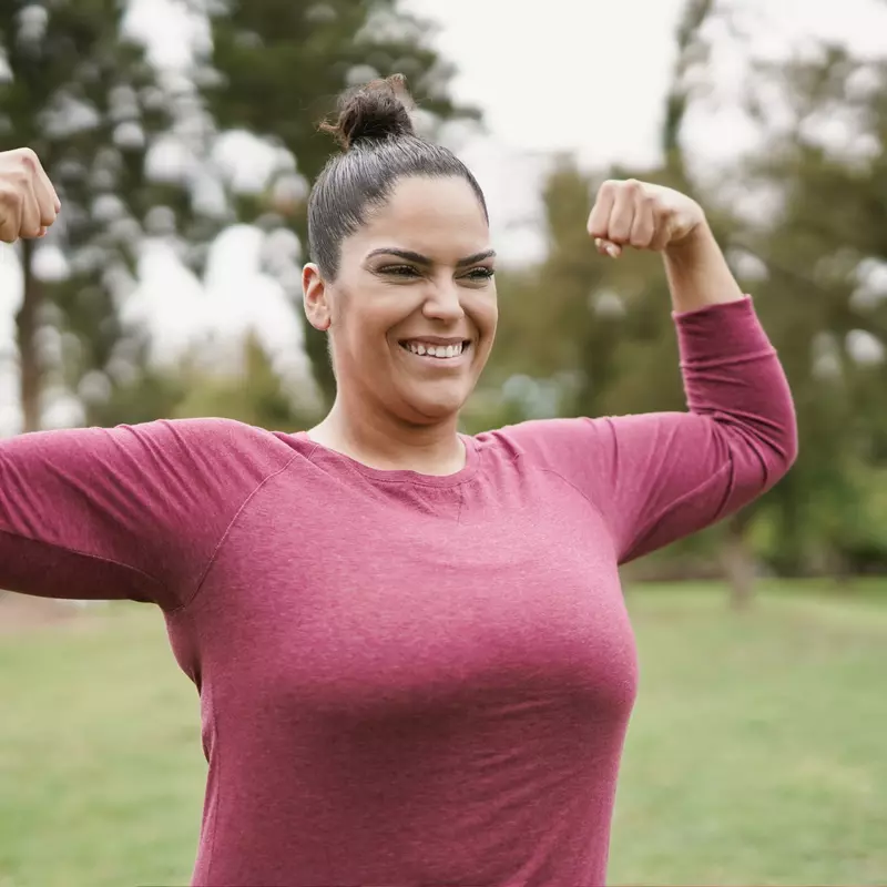 A Woman Flexes Her Muscles in a Park with a HUge Smile on Her Face.