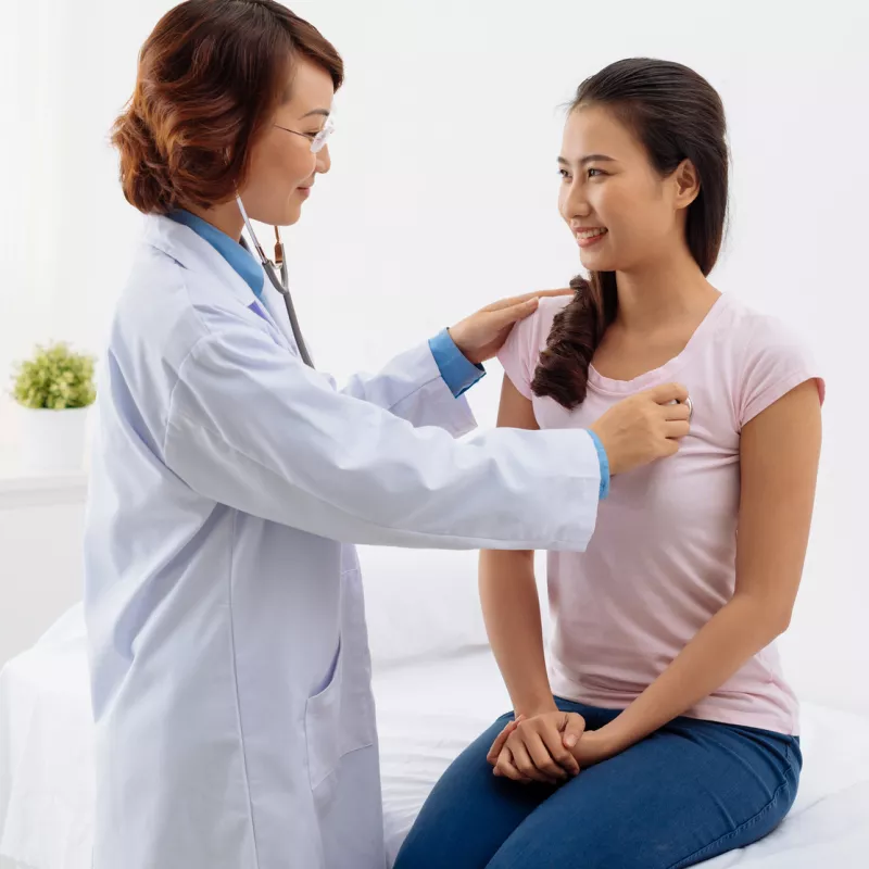 A woman is sitting on an exam table while a doctor listens to her heart.