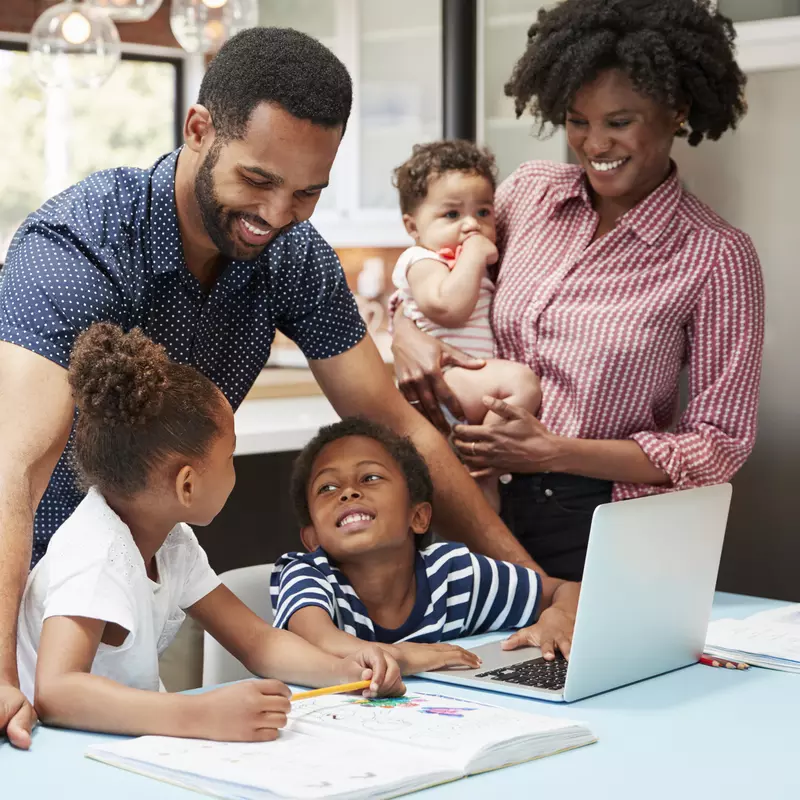 A family of five is in the kitchen, helping two of their children with their homework.