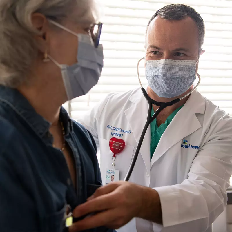 Doctor listening to patient's heartbeat with a stethoscope while both wear masks.