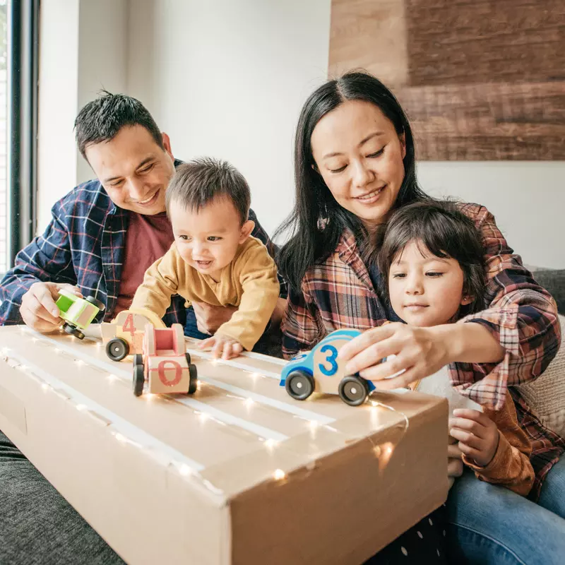 Parents and two children playing with wooden cars on top of a cardboard box while sitting on the couch at home.