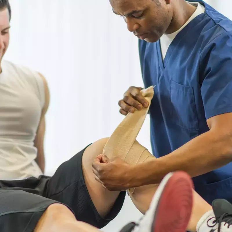 A physical therapist bandaging a male patient's left knee.